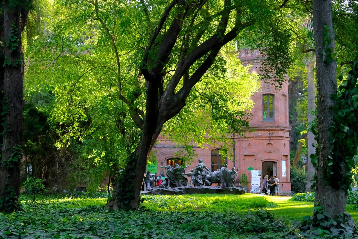 ต้นไม้ใบดกและไม้ใหญ่ล้อมอาคารอิฐแดงและประติมากรรมใน Jardín Botánico Carlos Thays เขต Palermo, Buenos Aires
