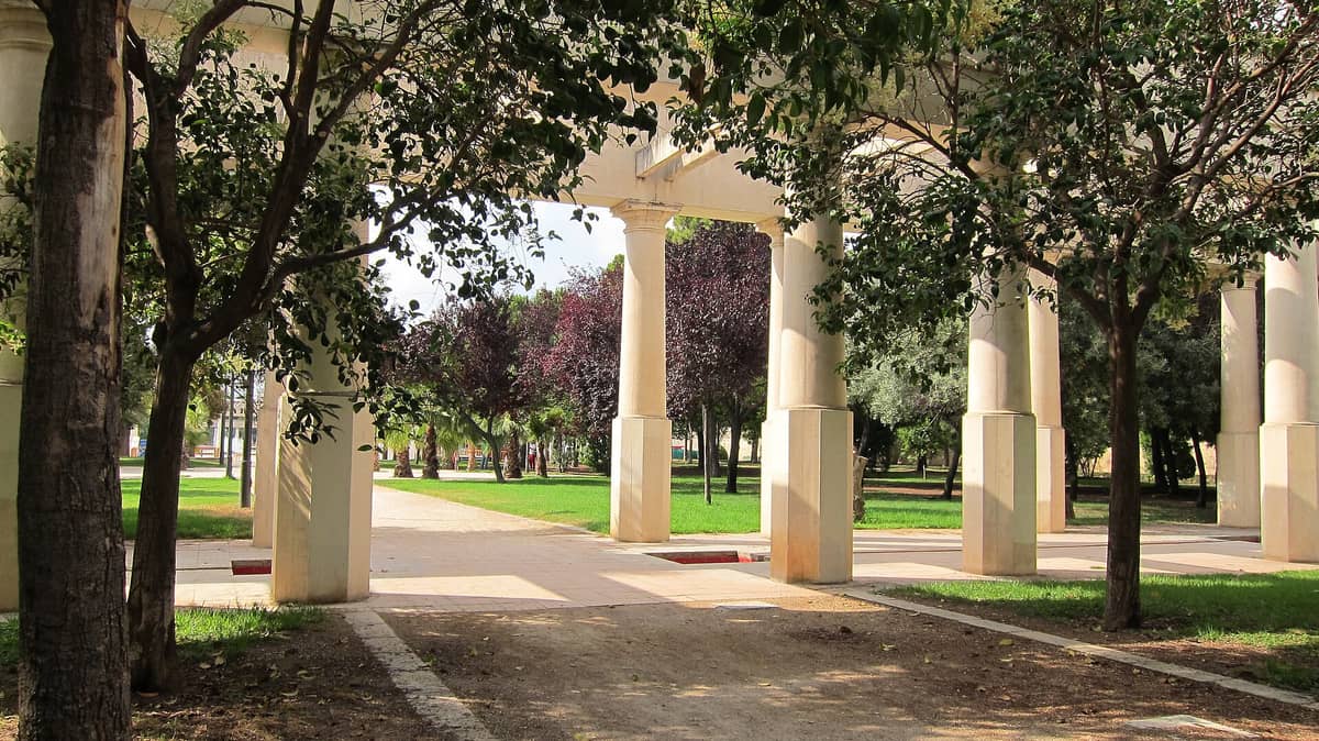 Un viale ombreggiato nel Jardín del Turia con colonne classiche, prato verde rigoglioso e alberi in un parco urbano.
