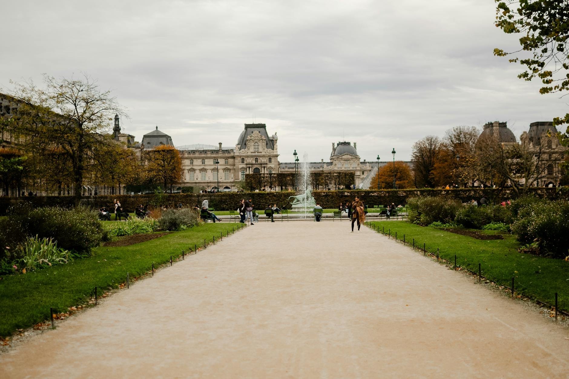 Ampio vialetto del Jardin des Tuileries con prati verdi, aiuole fiorite, persone che passeggiano, una fontana al centro e il Louvre sullo sfondo sotto un cielo nuvoloso.