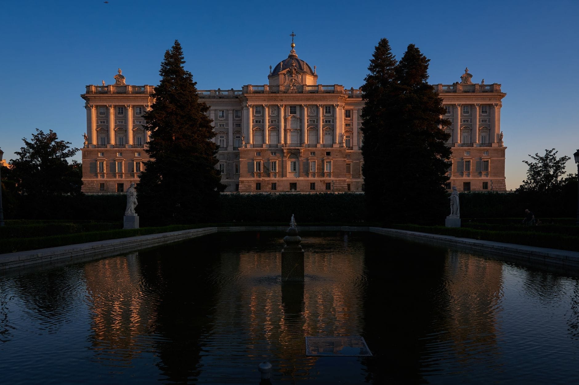 Vista del Palazzo Reale di Madrid con la sua magnifica facciata riflessa nello specchio d'acqua di un giardino formale al tramonto, incorniciata da statue e alberi alti.