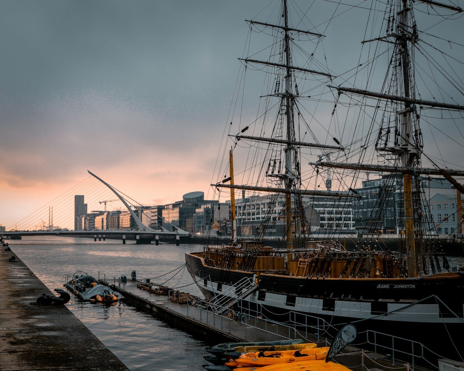 Il veliero Jeanie Johnston ormeggiato al Docklands di Dublino al tramonto, con lo skyline della città e il Ponte Samuel Beckett sullo sfondo.