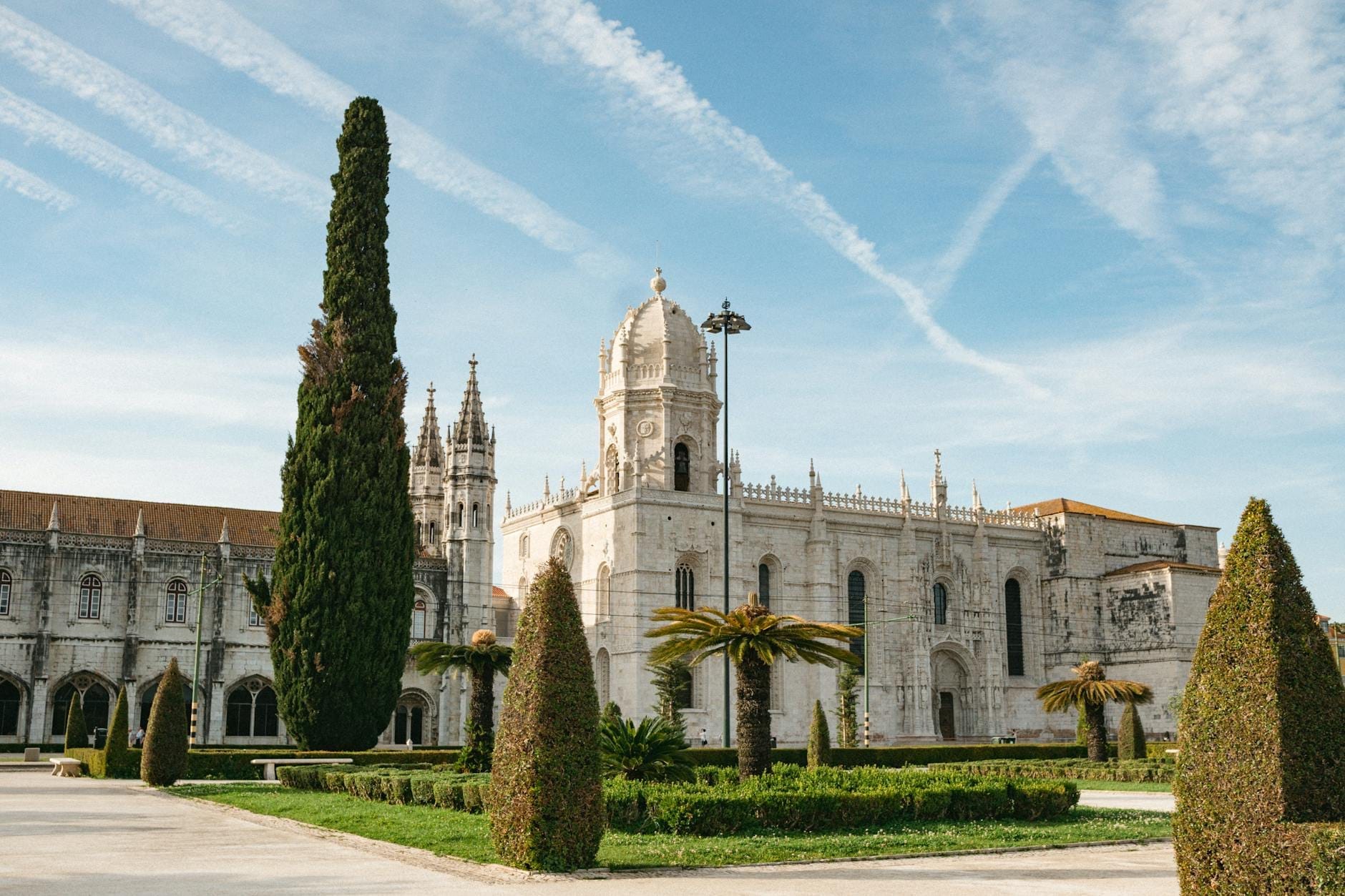 Vista panoramica del Monastero dos Jerónimos a Lisbona con la sua intricata architettura manuelina, la torre, i giardini e un cielo azzurro con leggere nuvole e scie di condensazione.