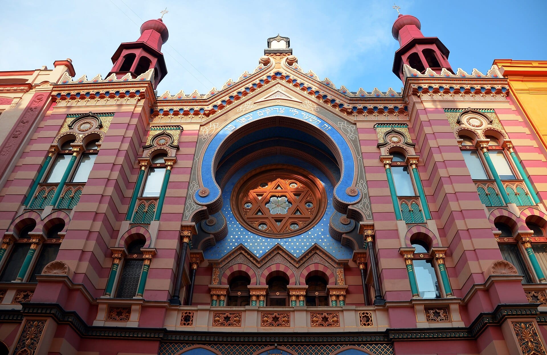 El exterior de la Sinagoga de Jerusalén en Praga presenta vibrantes paredes a rayas, arcos de estilo neoárabe, ventanas decorativas y coloridos detalles Art Nouveau bajo un cielo azul.