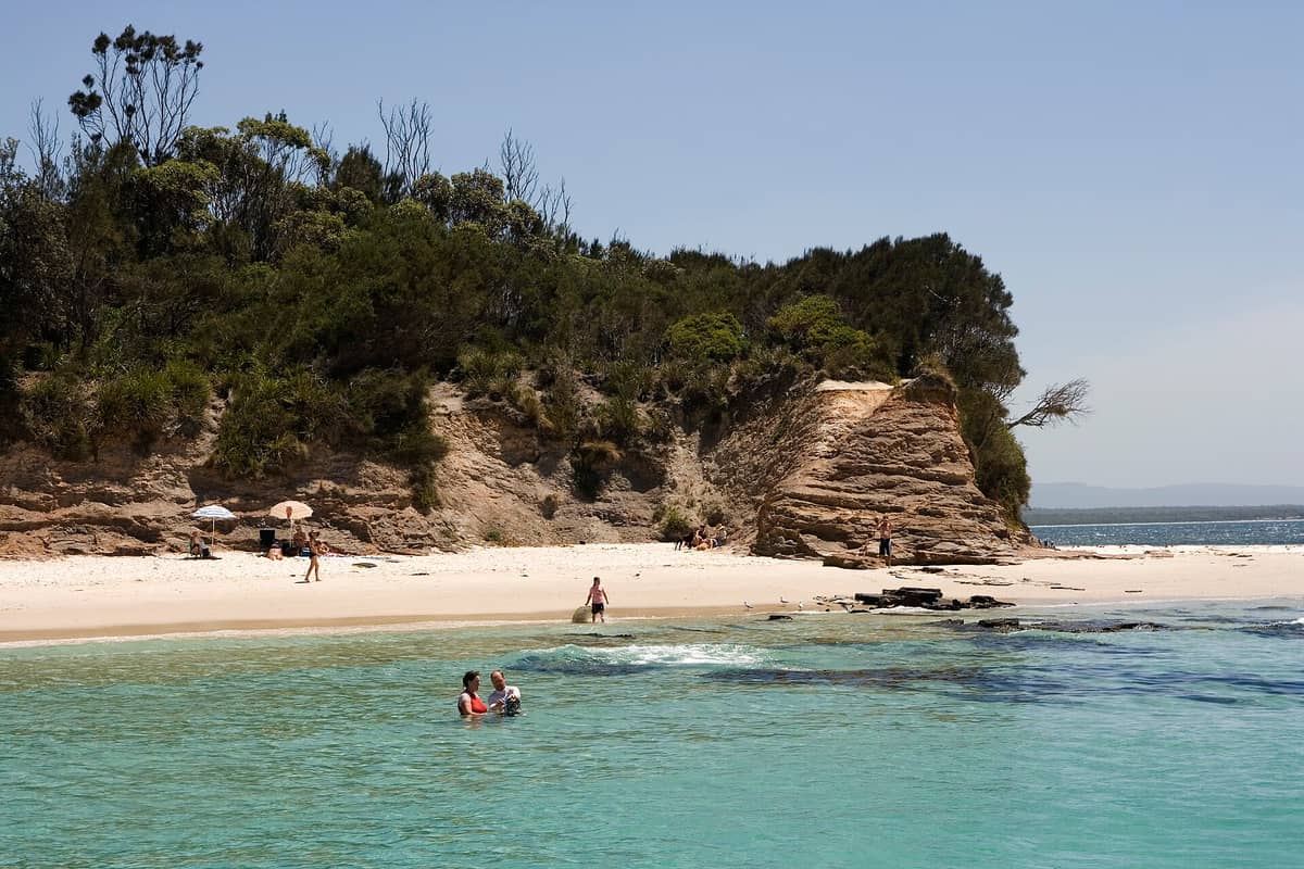 Menschen schwimmen und entspannen an einem weißen Sandstrand mit türkisklarem Wasser und buschigen Kaps unter einem wolkenlosen Himmel in Jervis Bay.