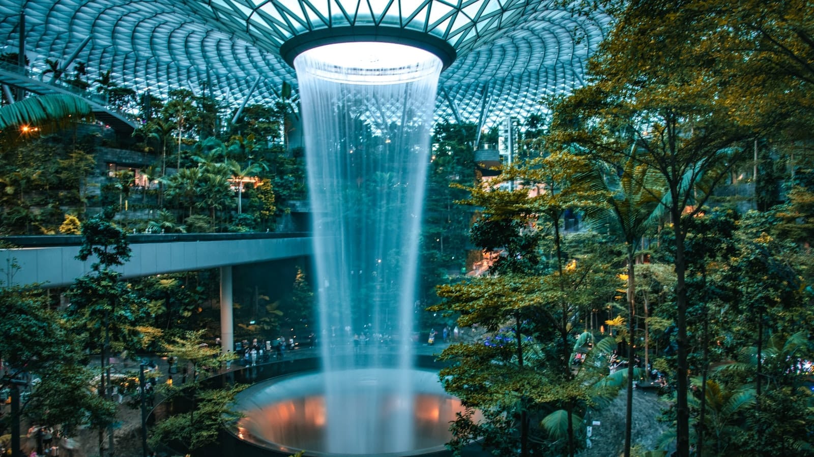 วิวกว้างของน้ำตก Rain Vortex ใน Jewel Changi Airport ไหลทะลุผ่านป่าในร่มเขียวชอุ่มภายใต้โดมกระจก พร้อมรถไฟฟ้า Skytrain และผู้เยี่ยมชมท่ามกลางต้นไม้เขียวขจี
