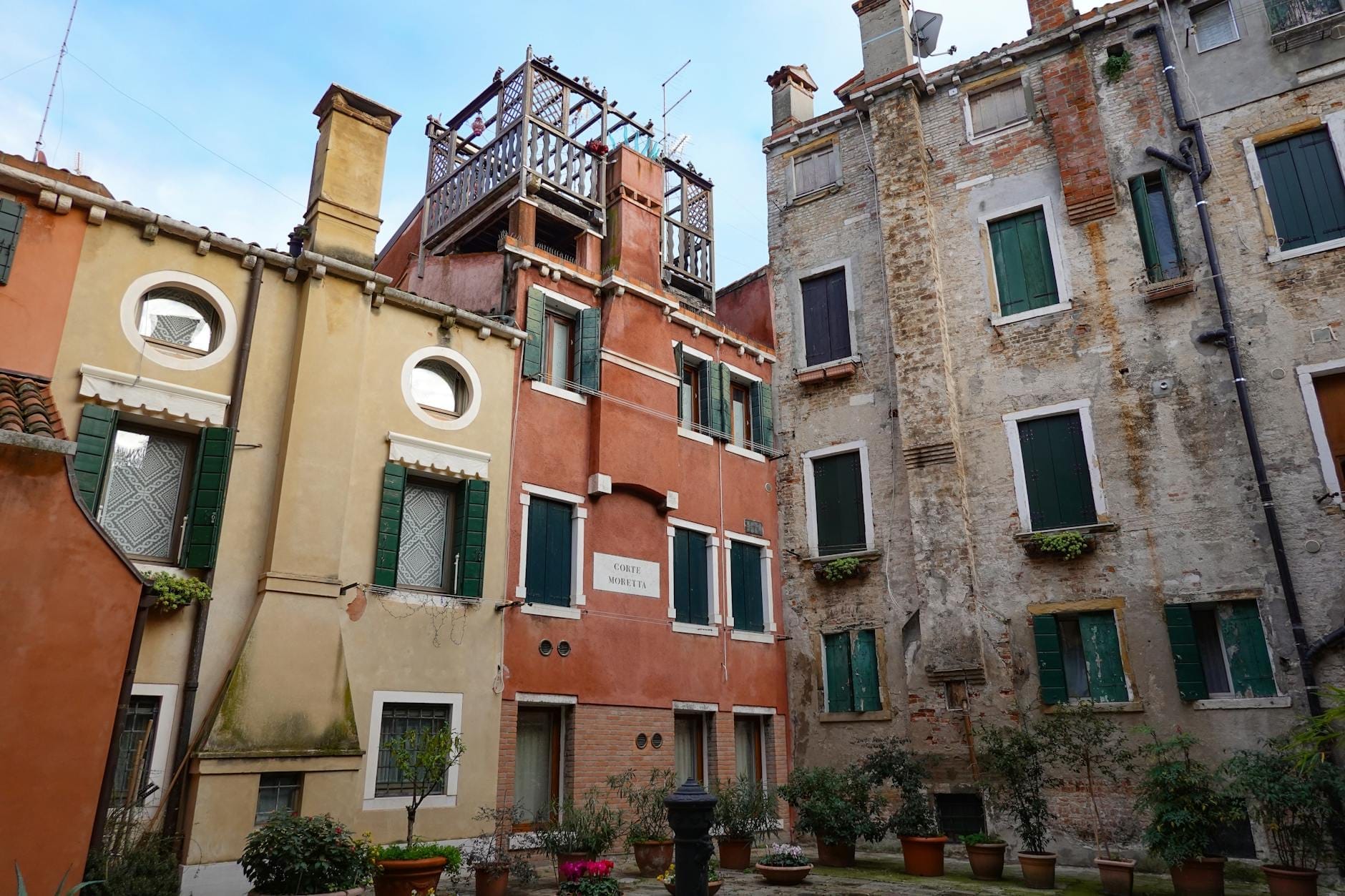 Colorful facades and weathered stone buildings with green shutters in Rome's Jewish Ghetto, surrounding a small courtyard with potted plants.