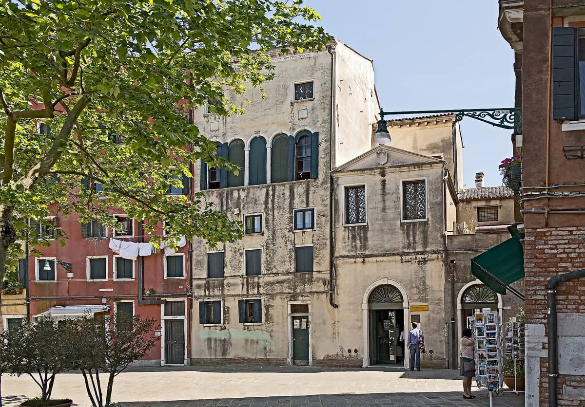 Blick auf den Hauptcampo im jüdischen Ghetto Venedigs mit historischen Gebäuden, dem Eingang des Jüdischen Museums und Bäumen im hellen Tageslicht.