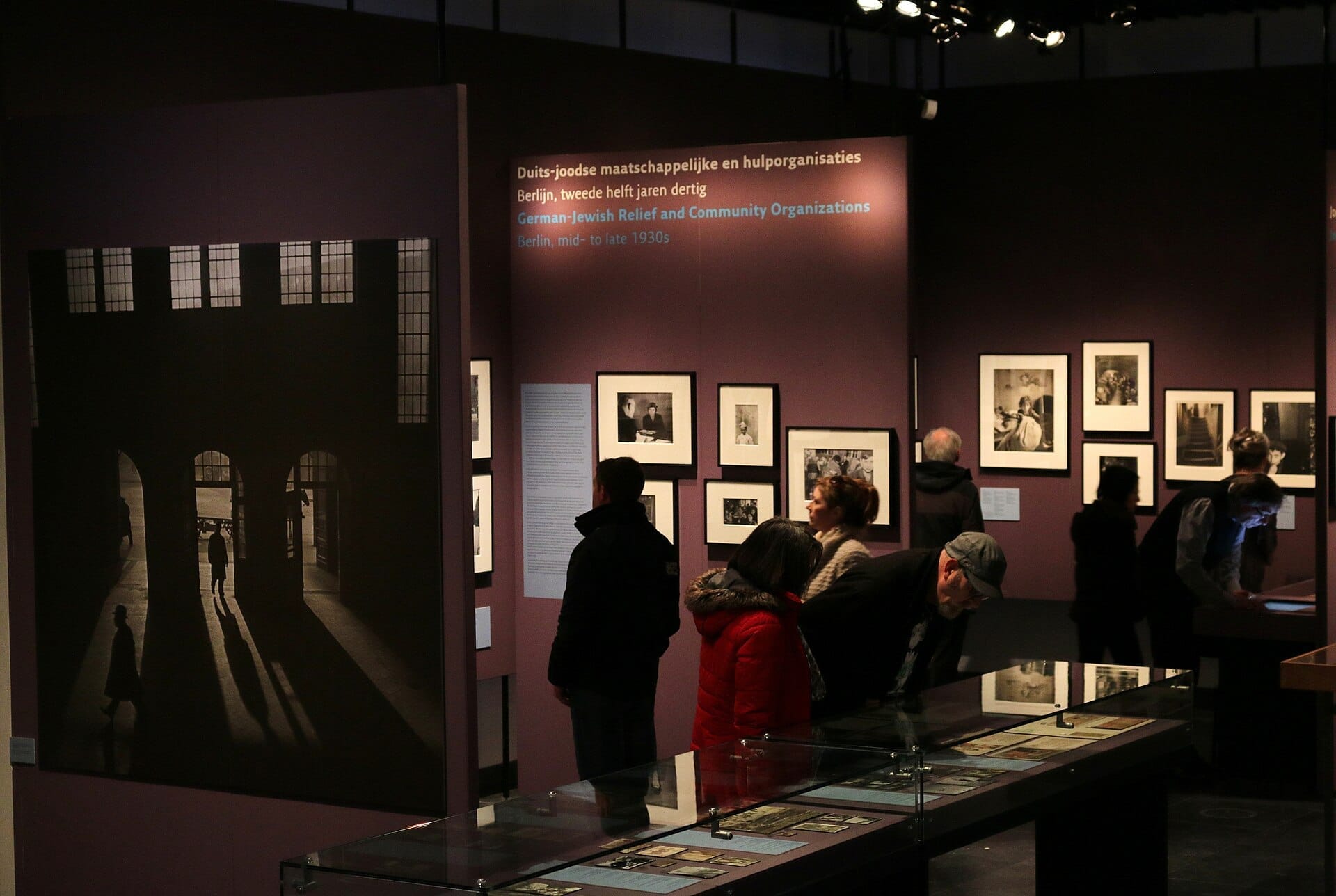 Visitantes examinan fotografías históricas y artefactos en una exposición tenuemente iluminada del Jewish Museum Amsterdam, con grandes imágenes en blanco y negro sobre paredes moradas.