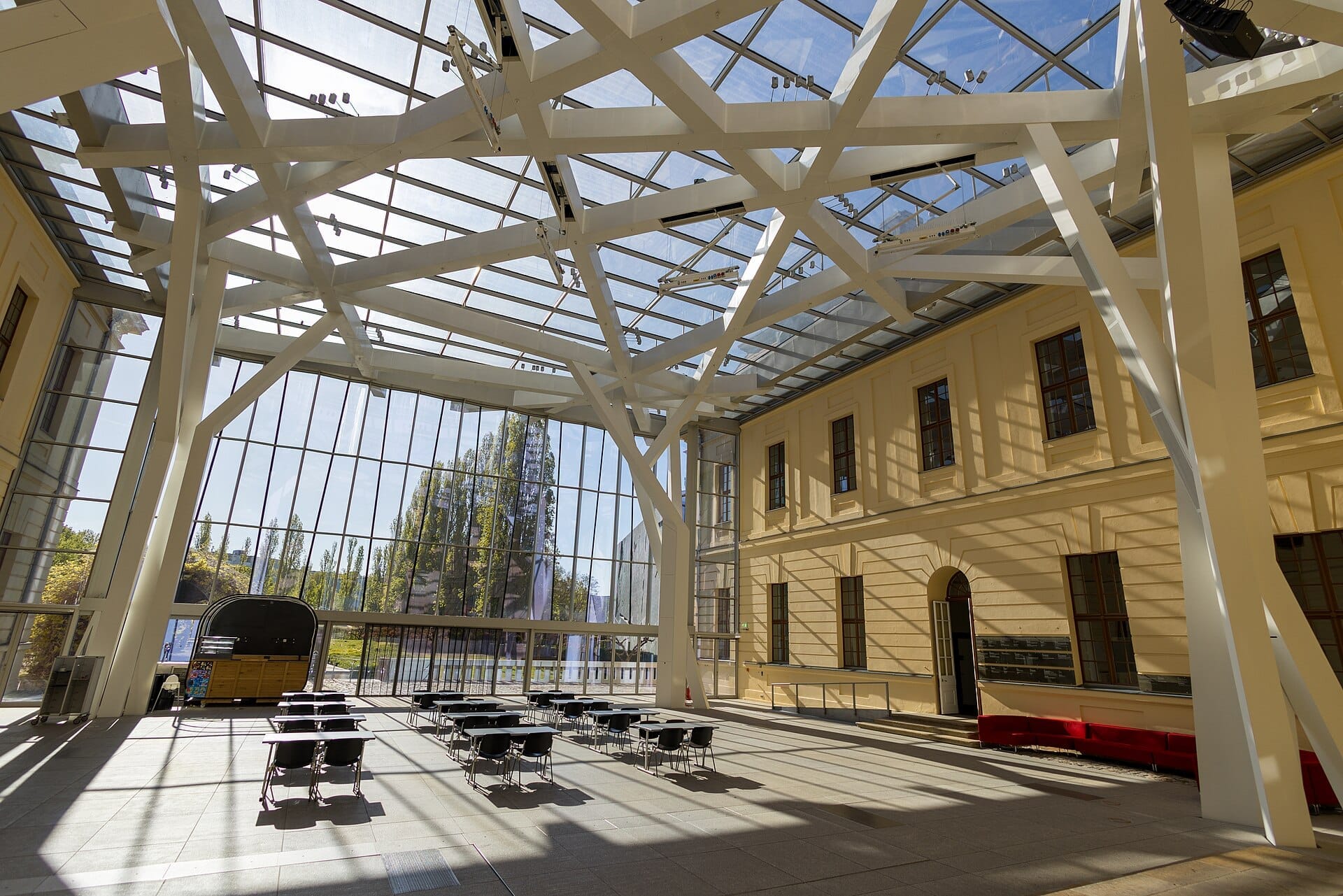 Ampio atrio all'interno del Jüdisches Museum Berlin con un moderno tetto in vetro, travi bianche geometriche e file di sedie nere sotto la luce naturale.