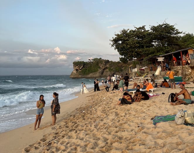 ผู้มาเยือนพักผ่อนบนหาดทรายขาวของหาดบาลังกันในจิมบารัน บาหลี ล้อมรอบด้วยหน้าผาหินปูนที่น่าทึ่ง วารุงริมหาด และคลื่นอ่อนภายใต้ท้องฟ้าแจ่มใส