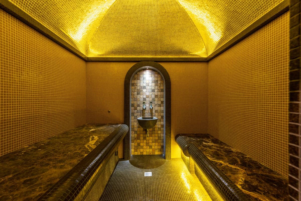 Sala de sauna com azulejos de mosaico, dois bancos de mármore e área central de lavagem sob iluminação amarela quente.