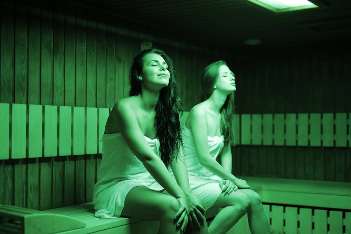 Duas mulheres com toalhas relaxando em uma sala de sauna com paredes e bancos de madeira, representando a experiência de um bathhouse ou jjimjilbang.