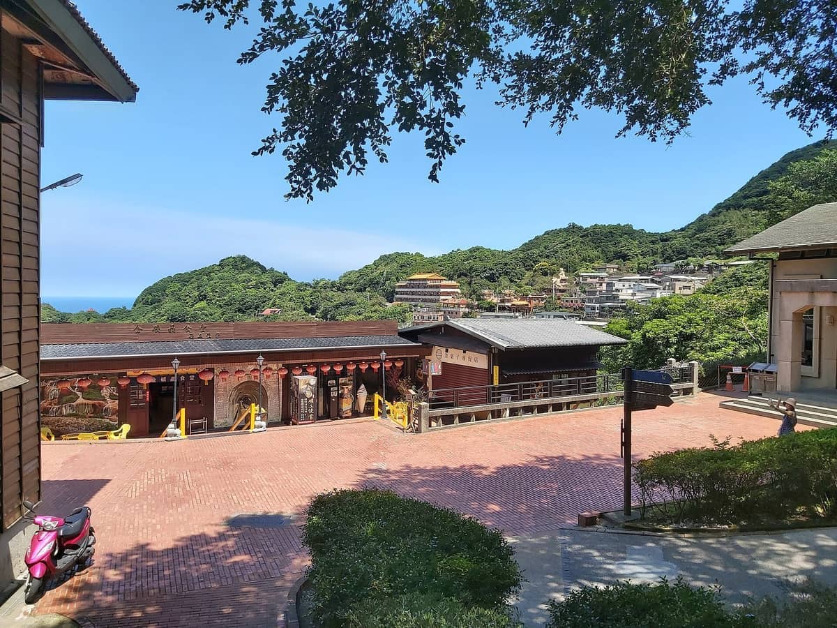 Blick auf die Gebäude des Jinguashi Goldmuseums mit rotem Backsteinplatz, üppigen grünen Berghängen, verstreuten Häusern und klarem blauem Himmel in Ruifang, Taiwan.