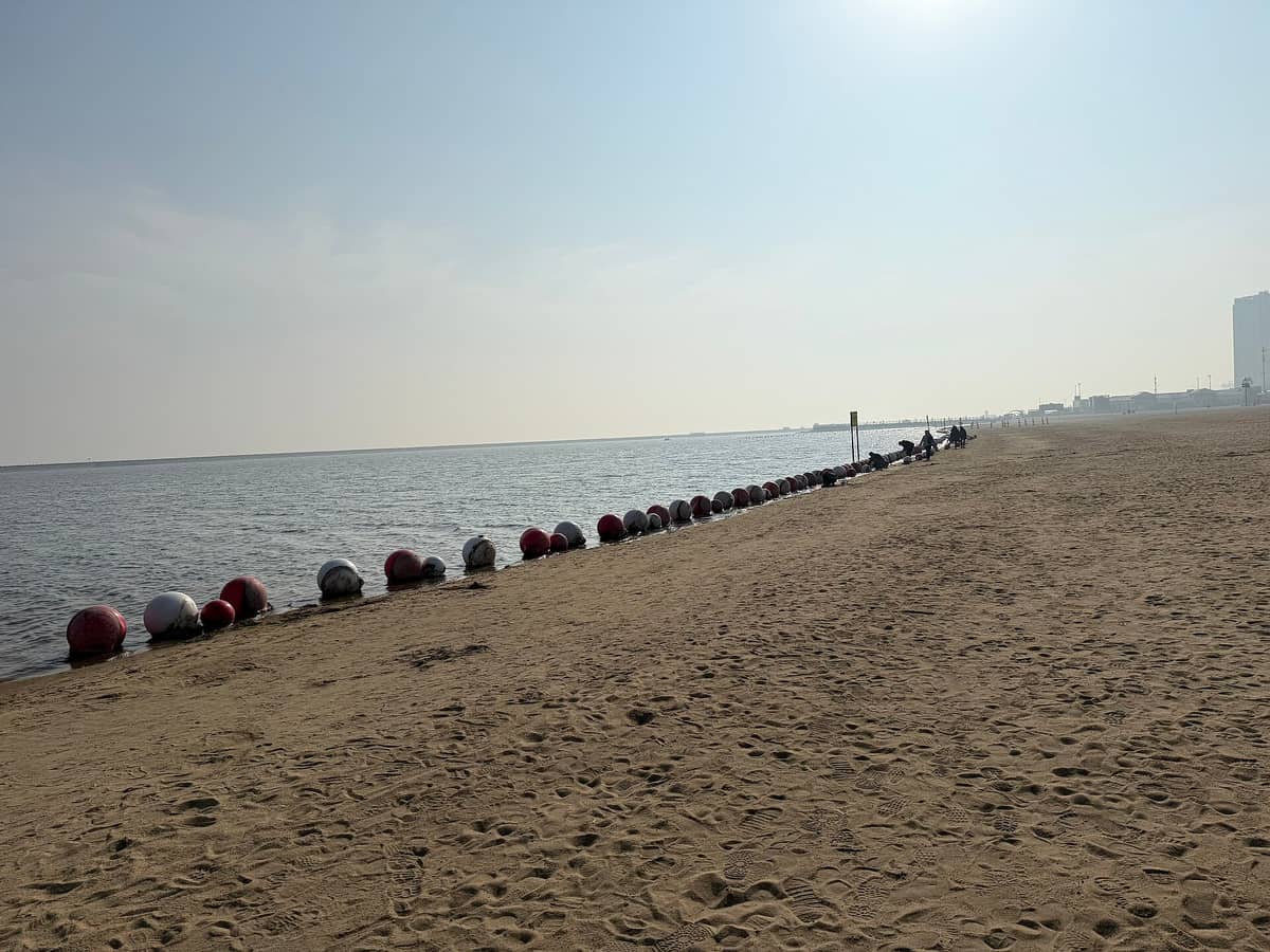 Weitläufiger Sandstrand des Jinshan City Beach in Shanghai mit rot-weißen Bojen, die eine Schwimmzone entlang der ruhigen Küste unter einem diesigen Himmel markieren.