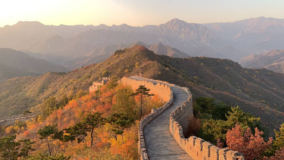 Panoramablick auf die Jinshanling-Mauer, die sich im Herbst über Bergrücken schlängelt – mit goldenem Laub, dramatischen Gipfeln und steinernen Wachtürmen im sanften Abendlicht.