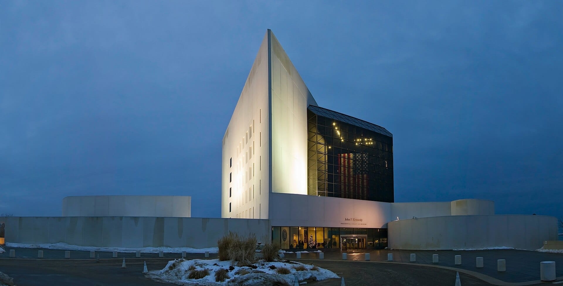 La John F. Kennedy Presidential Library and Museum al crepuscolo, con architettura moderna illuminata, facciata in vetro e chiazze di neve a terra.