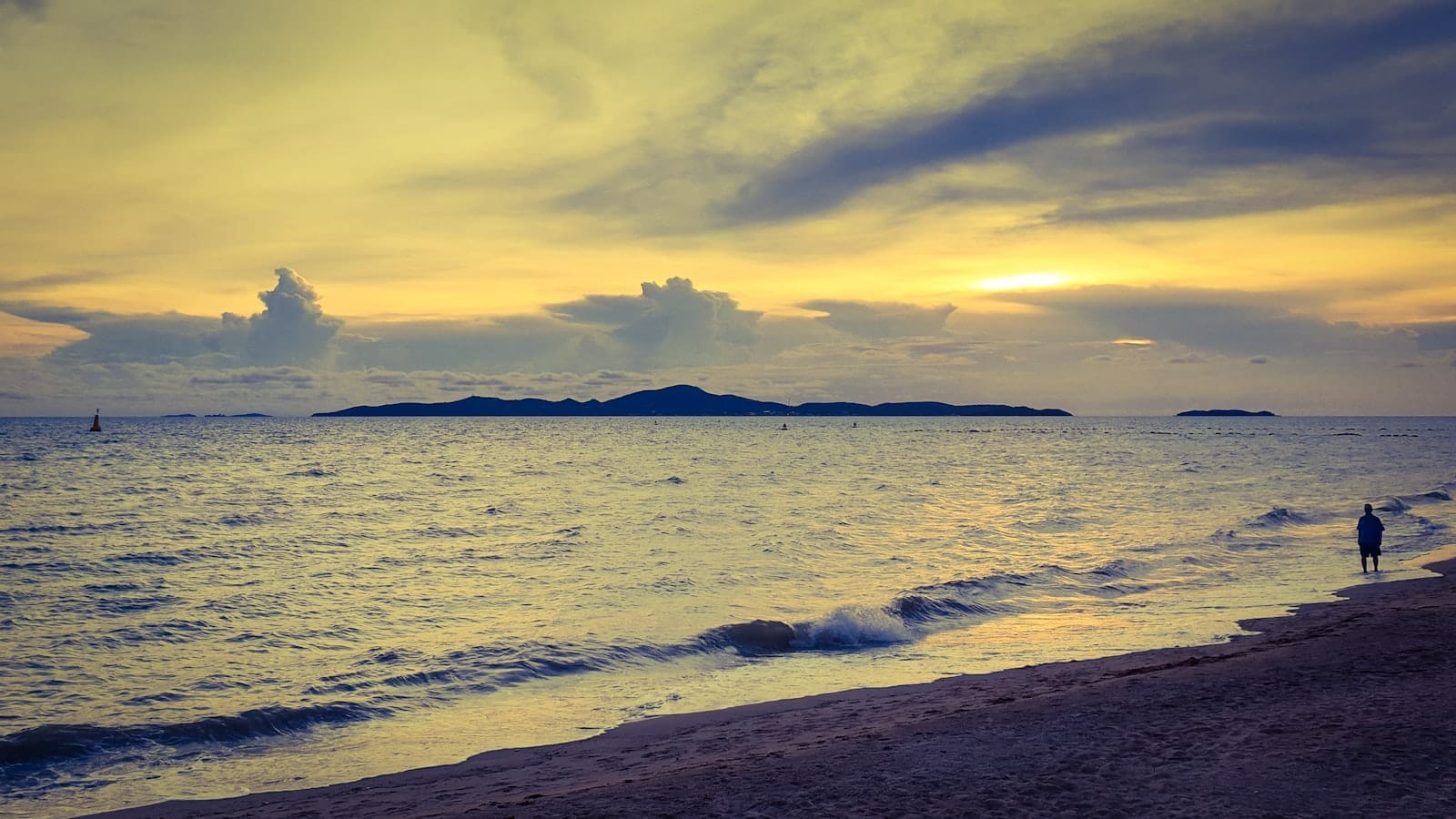 La riva tranquilla di Jomtien Beach al tramonto, con onde leggere, sabbia dorata e una figura solitaria in lontananza che cammina lungo il bagnasciuga.