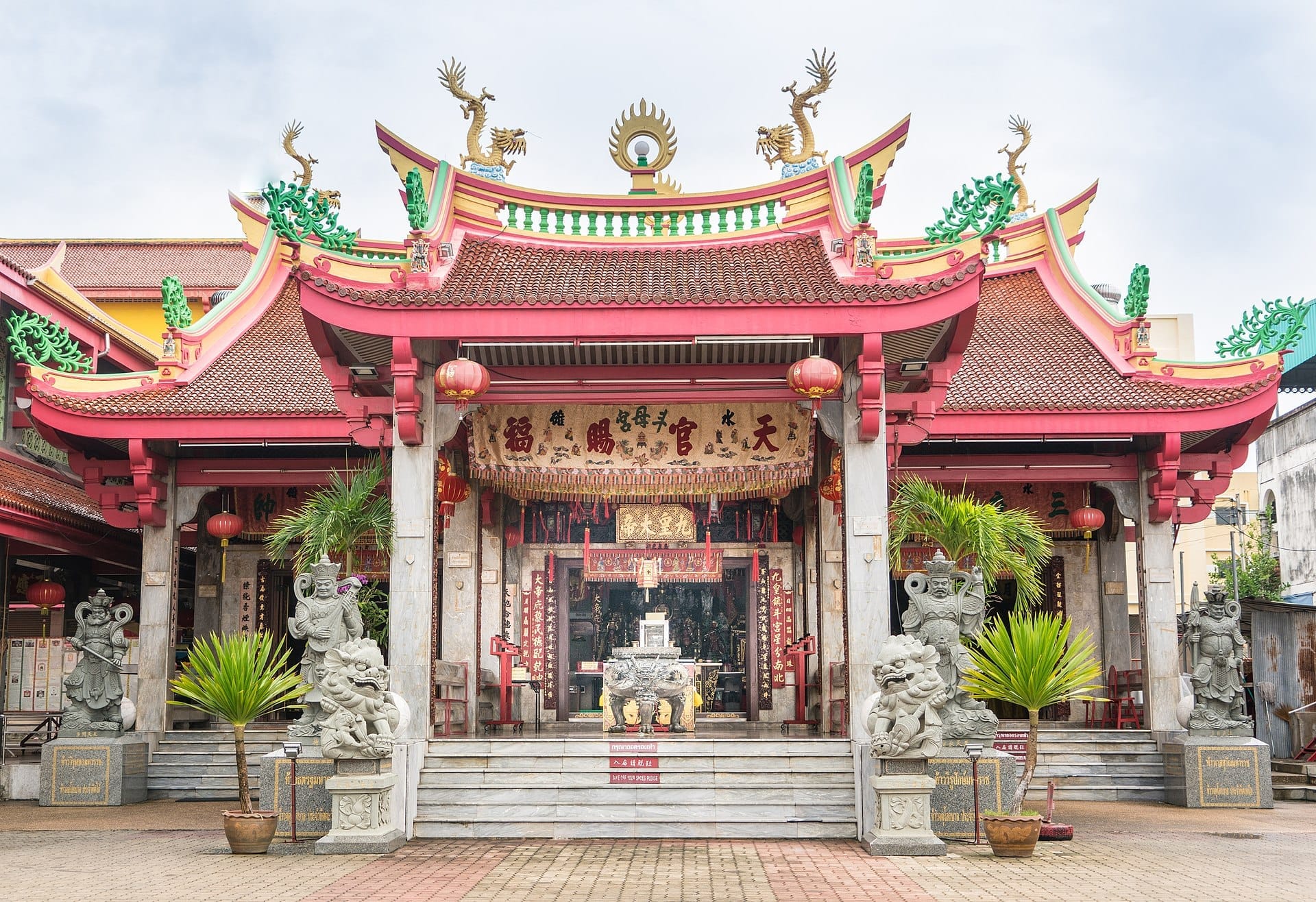Front view of Jui Tui Shrine in Phuket, featuring ornate red rooftops, stone statues, Chinese lanterns, and decorative banners under bright daylight.
