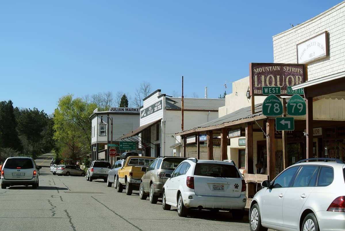 Die historische Main Street in Julian, Kalifornien, mit geparkten Autos, altmodischen Ladengeschäften und grünen Bergen im Hintergrund an einem sonnigen Tag.