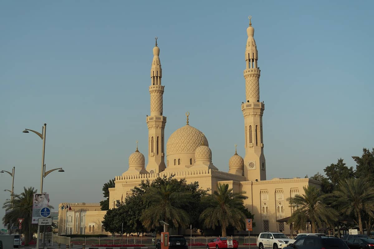 Weitwinkelperspektive der Jumeirah-Moschee in Dubai mit weißer Steinfassade, zentraler Kuppel, zwei Minaretten, Palmen und geparkten Autos unter klarem Himmel.
