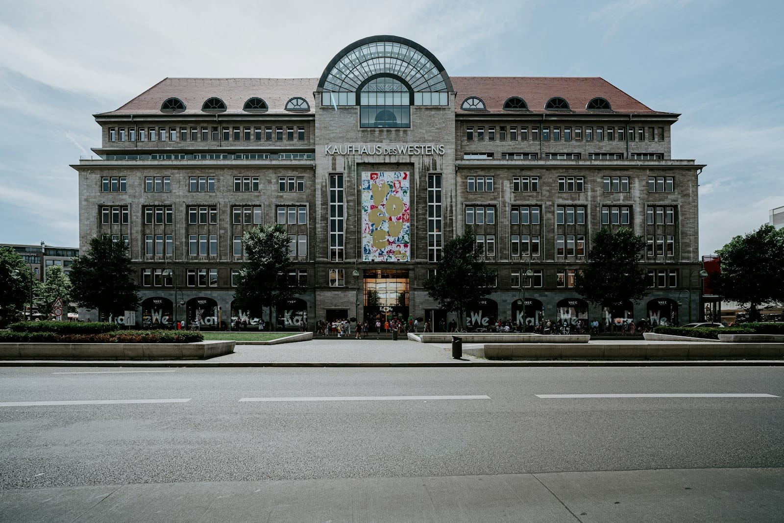 Vista frontale dello storico grande magazzino KaDeWe a Berlino, con la sua maestosa facciata, alberi e un colorato striscione all'ingresso sotto un cielo nuvoloso.