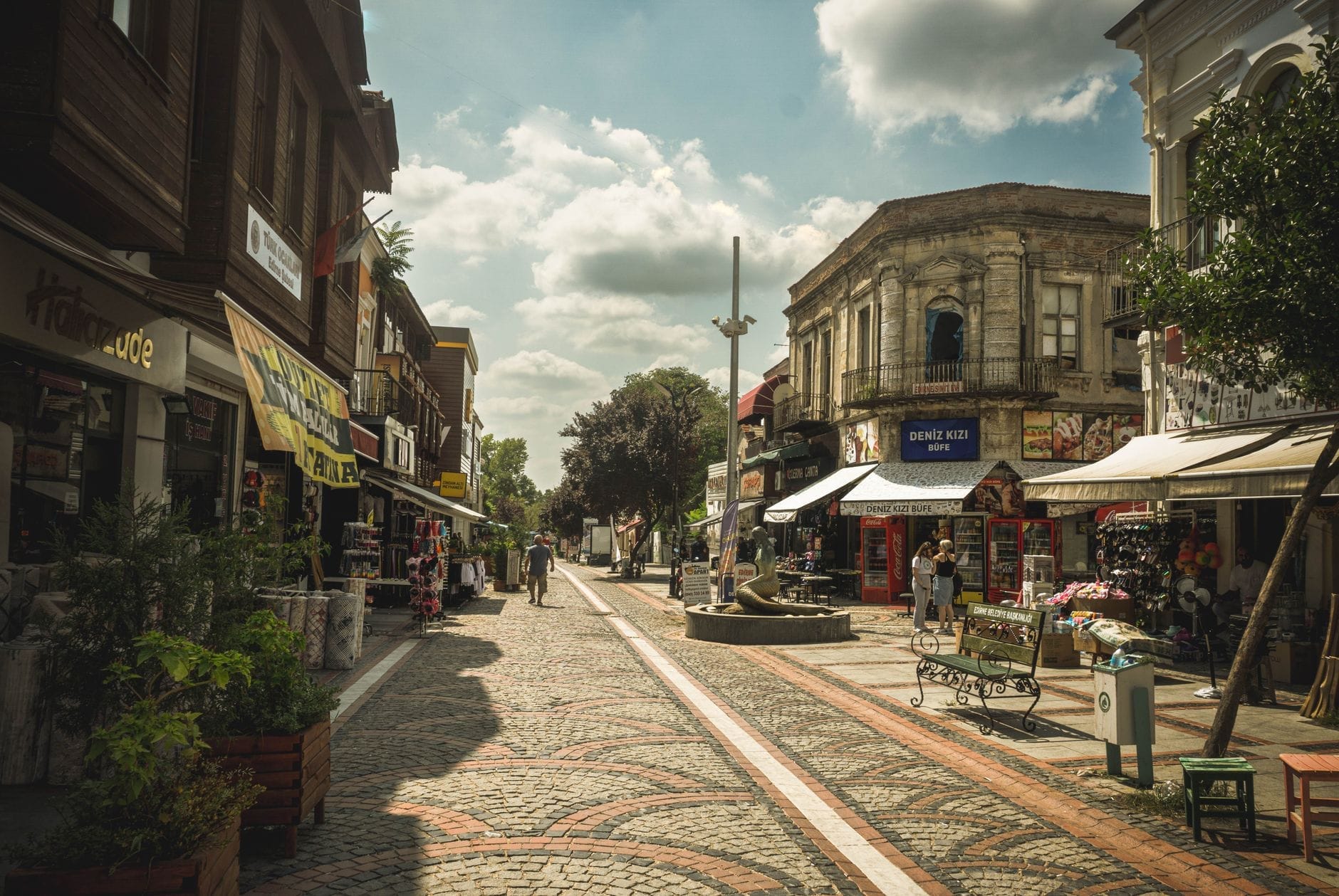 Una strada pedonale assolata nel Quartiere del Mercato di Kadıköy, fiancheggiata da negozi, caffè ed edifici storici, con un'atmosfera vivace ma rilassata.