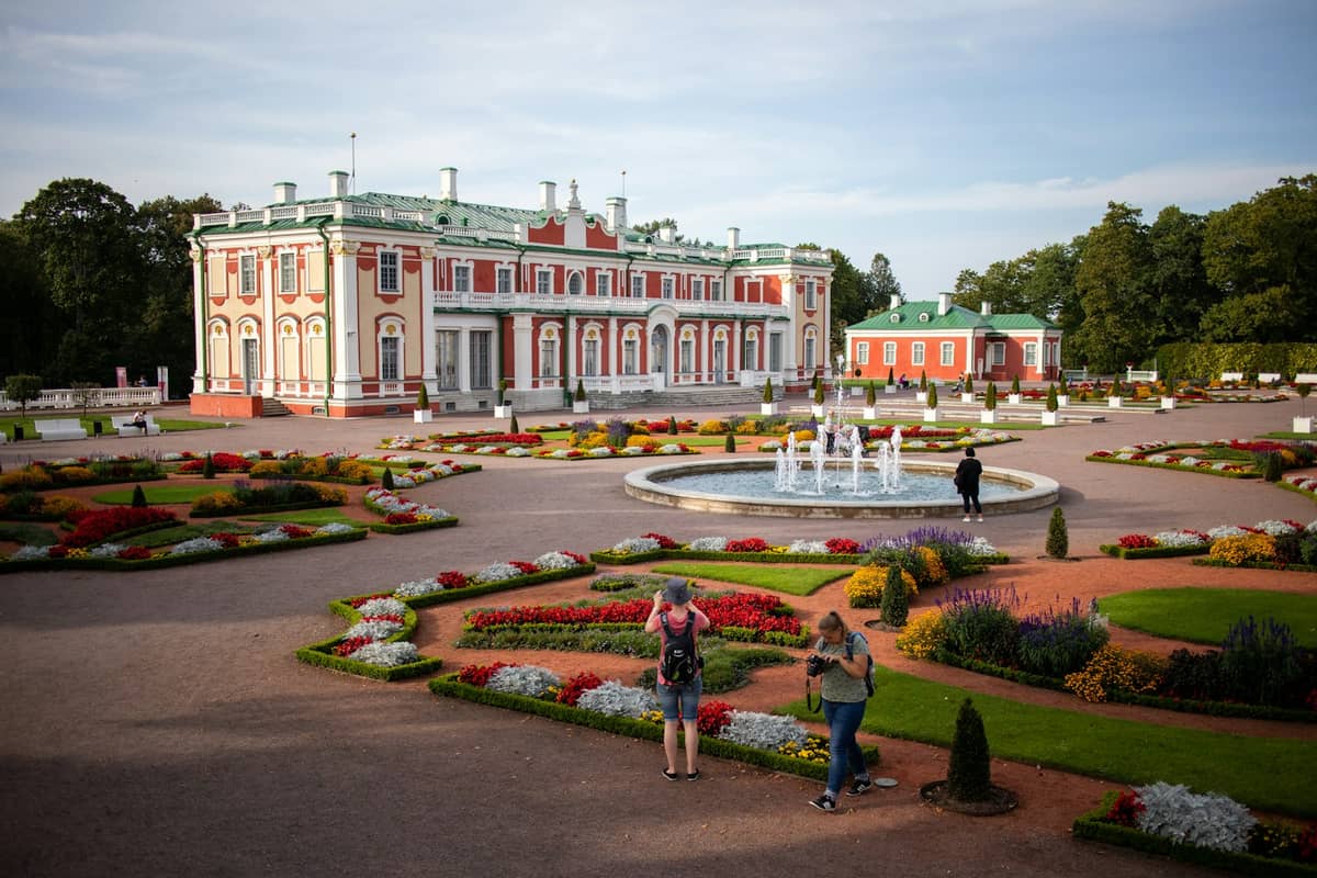 Der Kadriorger Palast in Tallinn mit seiner reich verzierten Barockfassade, Gartenanlagen, bunten Blumenbeeten, einem zentralen Springbrunnen und Besuchern im Vordergrund.
