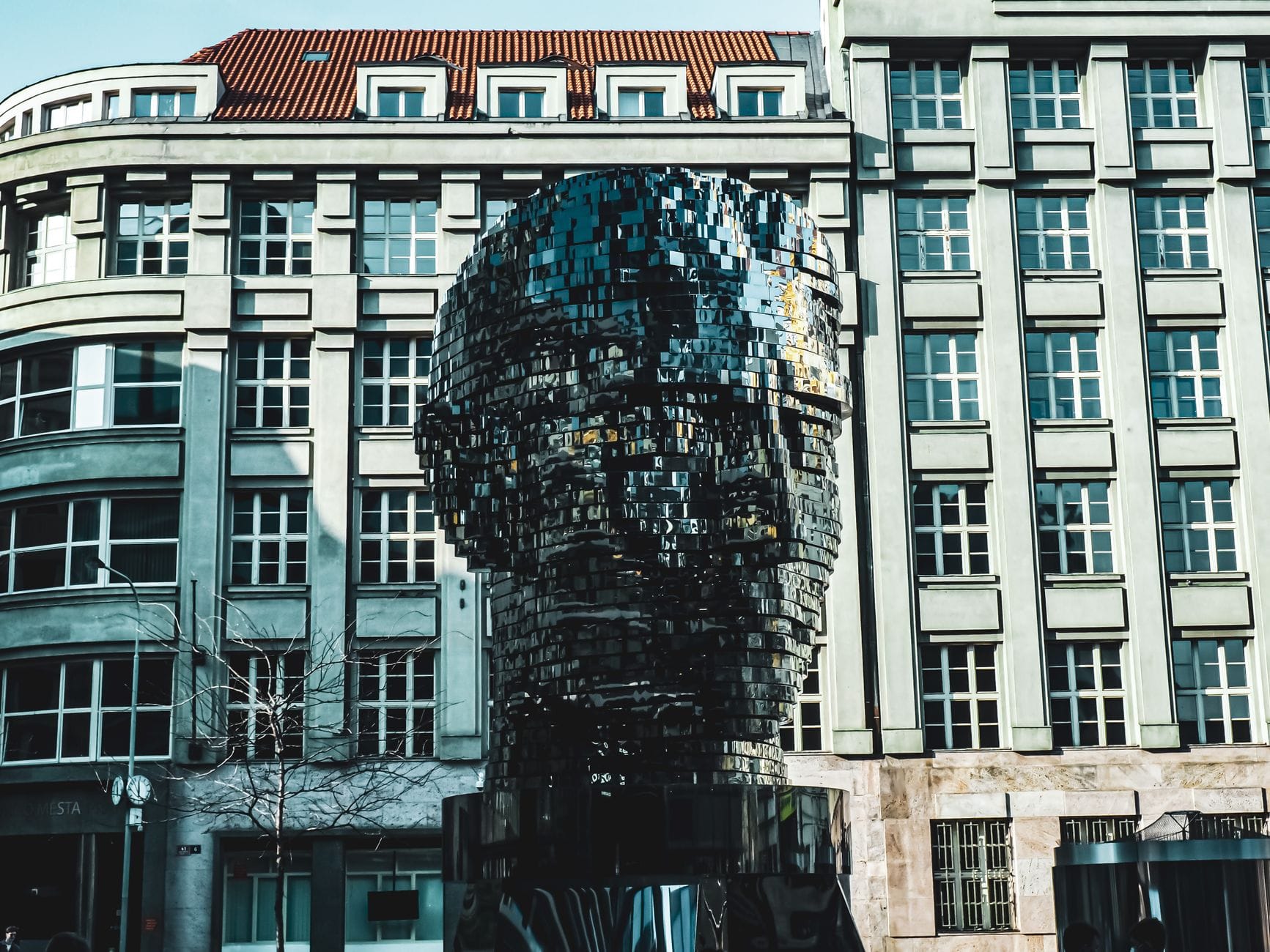 La scultura della Testa Rotante di Kafka a Praga, con gli strati in acciaio inox specchiato che riflettono gli edifici circostanti, fotografata di giorno davanti al centro commerciale Quadrio.