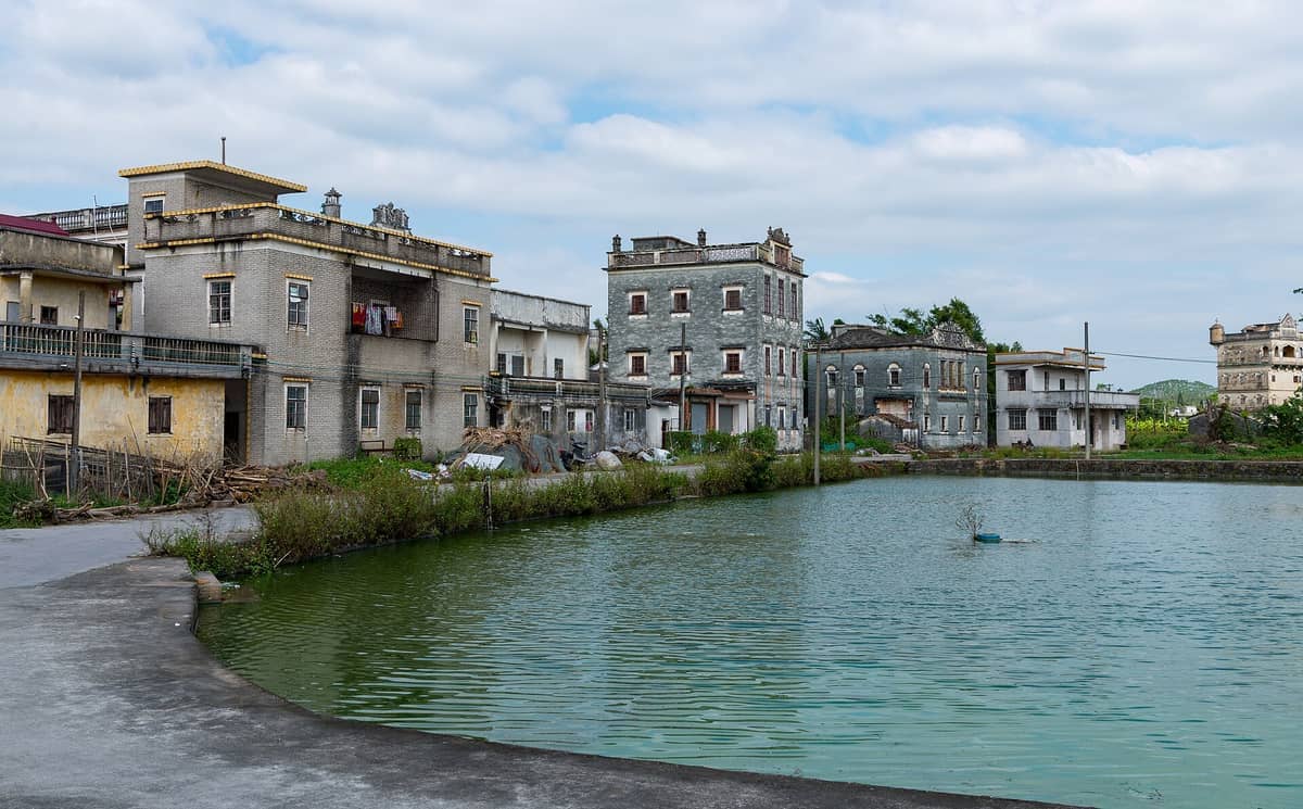 Historic Kaiping Diaolou towers and village homes reflected in a tranquil pond under a partly cloudy sky in Guangdong, China.