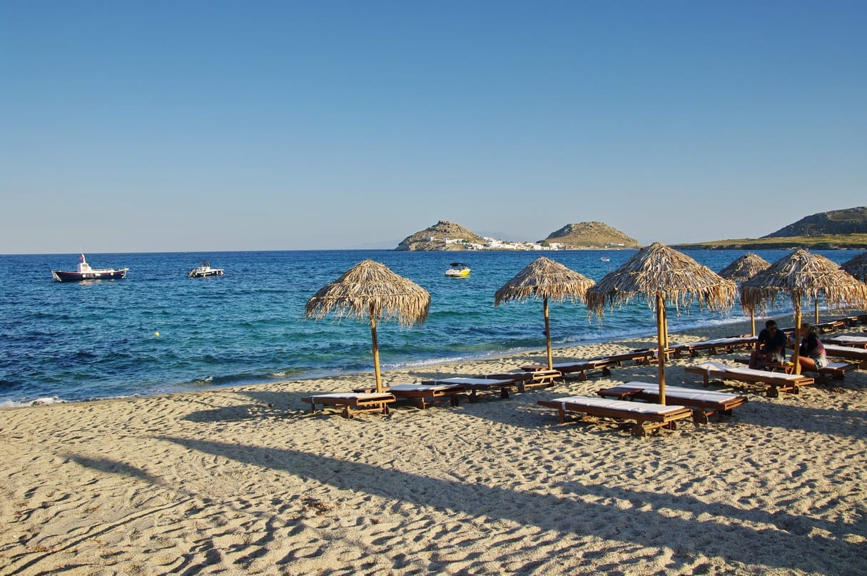 Playa Kalafatis de arena en Mykonos con sombrillas de paja, tumbonas, mar azul cristalino y lejanas colinas rocosas bajo un cielo soleado.