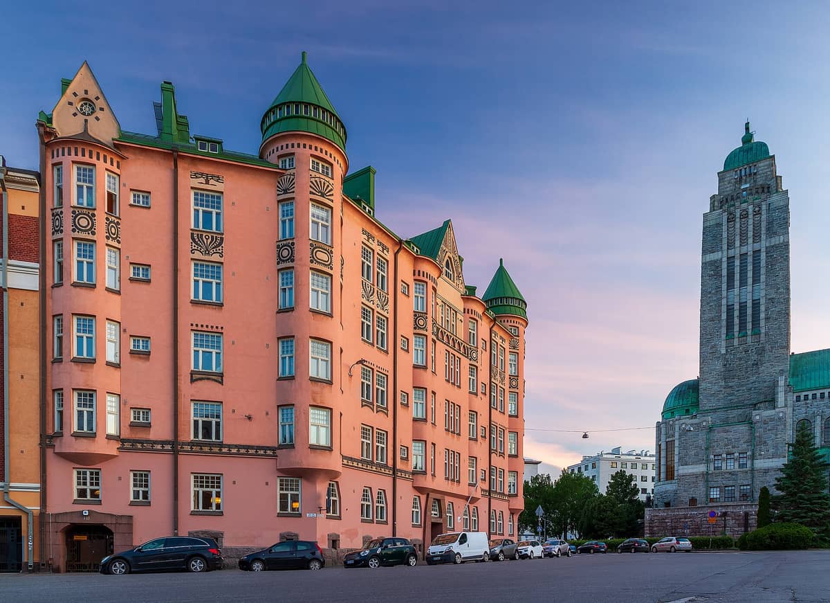 Prédio art nouveau e Igreja de Kallio no bairro de Kallio, em Helsinque, ao pôr do sol, com céu rosado e rua em primeiro plano.