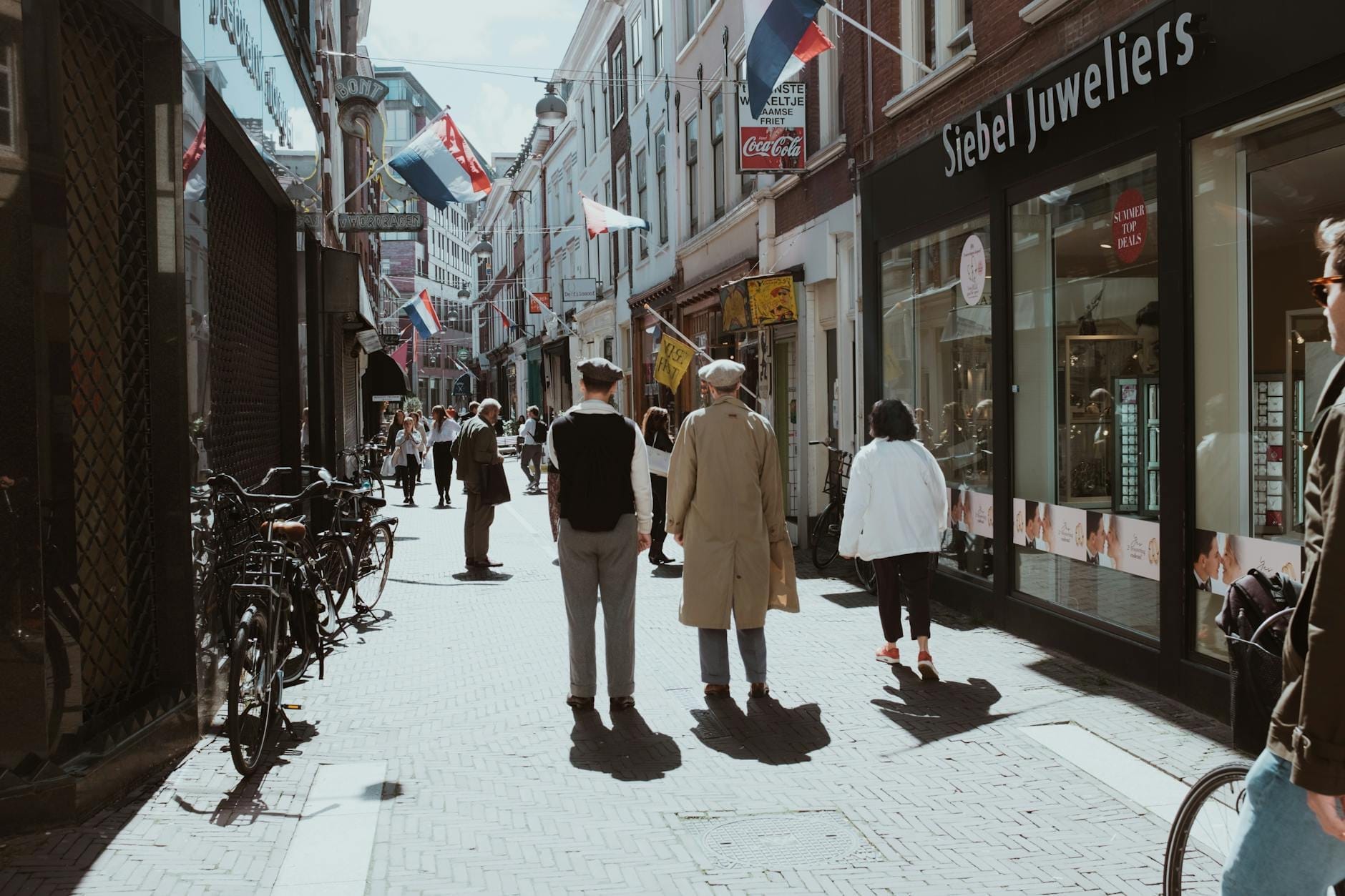 Peatones caminando por Kalverstraat en Ámsterdam, con bicicletas aparcadas en la acera, banderas holandesas y tiendas a lo largo de la soleada calle.