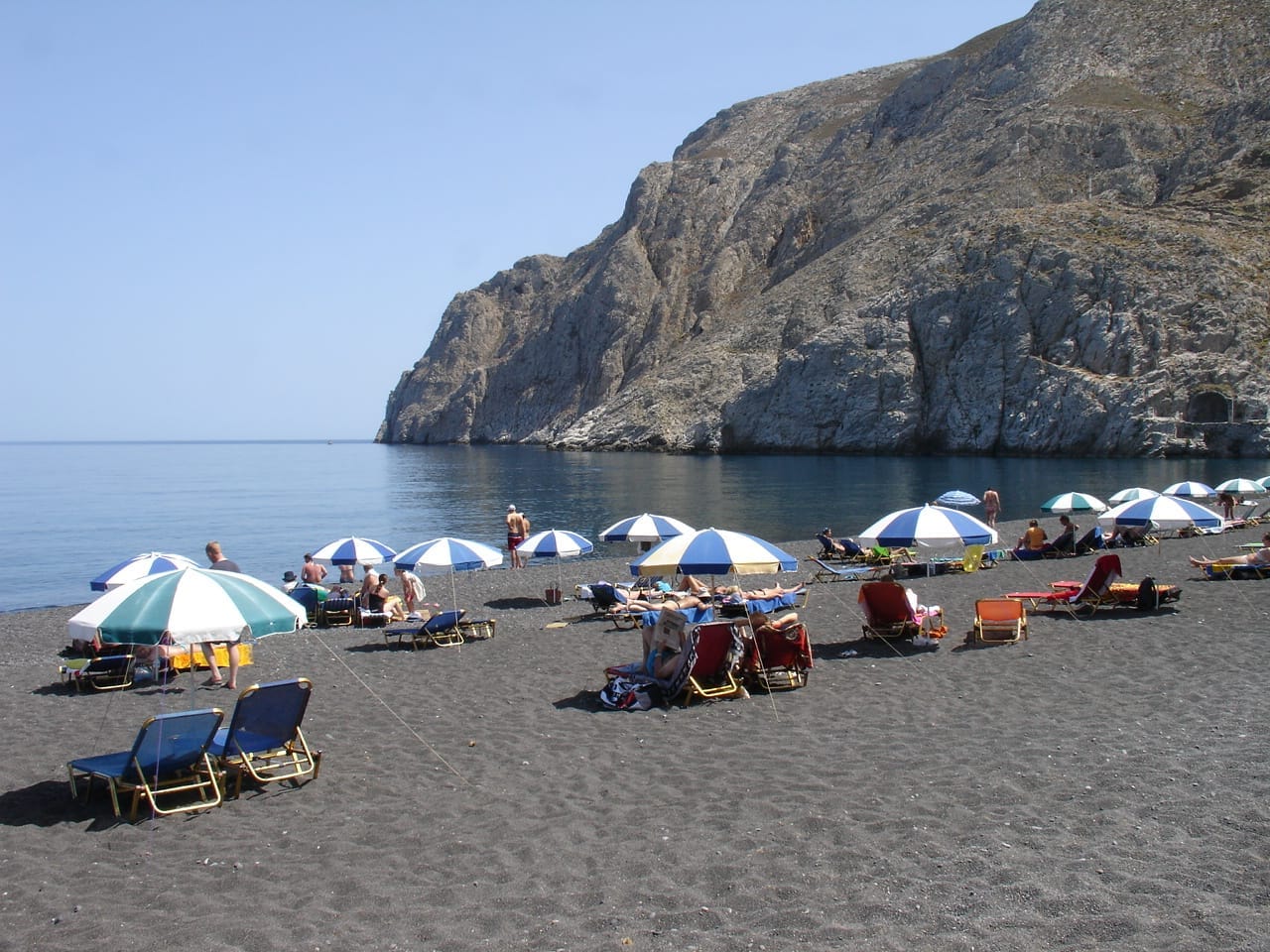 หาด Kamari ใน Santorini พร้อมเก้าอี้อาบแดด ร่มชายหาด ผู้คนที่พักผ่อน และหน้าผาหินที่น่าตื่นตาตื่นใจภายใต้ท้องฟ้าสีฟ้าแจ่มใส