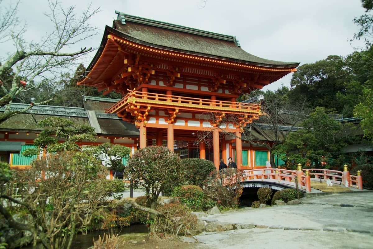Vibrant red torii gate and wooden bridge at Kamigamo Shrine, surrounded by manicured greenery and traditional architecture under a cloudy sky.