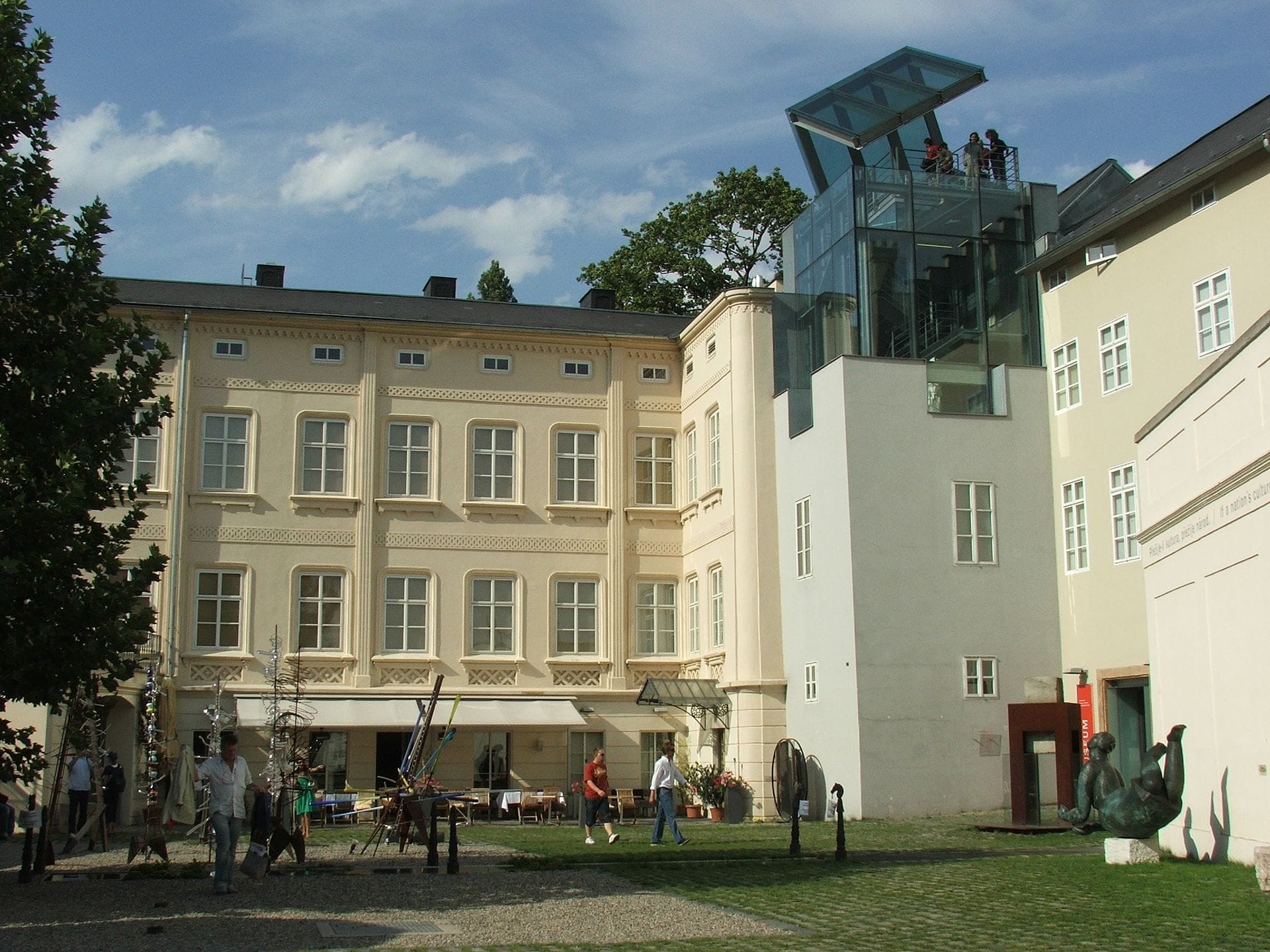L'esterno del Museum Kampa presenta un edificio storico restaurato color crema, un ascensore in vetro moderno, sculture all'aperto e visitatori che passeggiano nel cortile in una giornata di sole.
