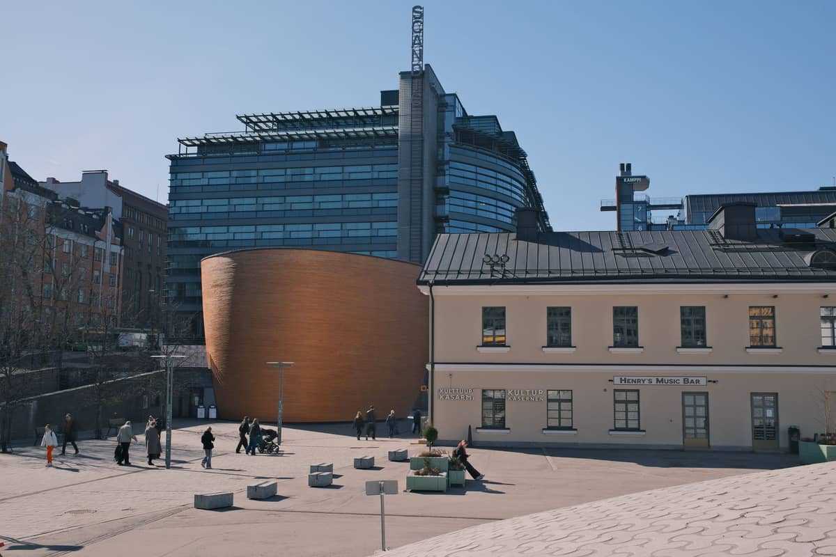 A Capela do Silêncio de Kamppi, uma estrutura curvilínea de madeira, na Praça Narinkka em Helsinque, com pessoas caminhando pela praça aberta em um dia ensolarado.