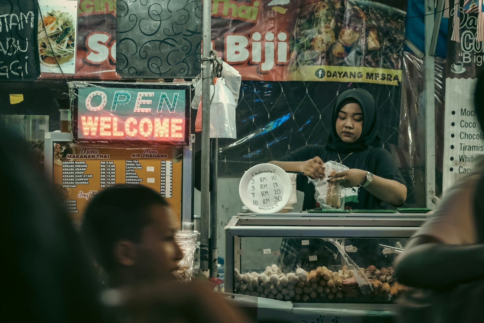 Traveler's view of Kampung Baru street market, highlighting a vendor packaging food as visitors browse local Malay delicacies.