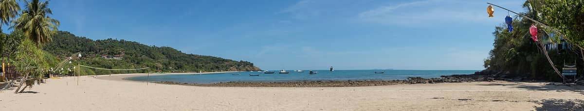 Vista panorámica de la bahía de Kantiang en Koh Lanta: arena fina, mar turquesa, olas suaves, colinas boscosas, barcas de pesca y una orilla tranquila y poco concurrida bajo un cielo azul.