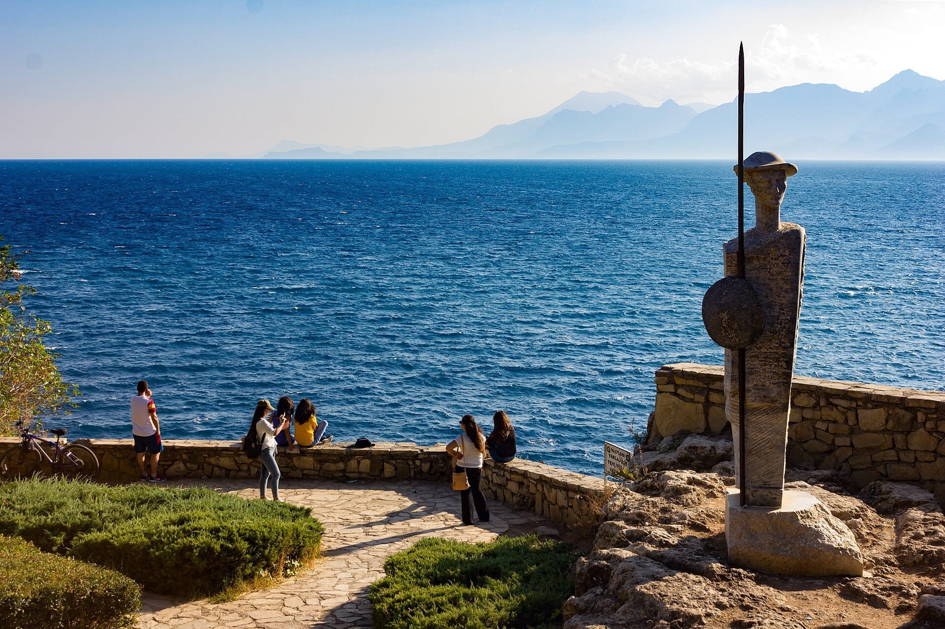 Vista panoramica dal Parco Karaalioğlu che mostra un sentiero in pietra, una statua sul lungomare e visitatori che si affacciano sul luminoso Mar Mediterraneo azzurro.