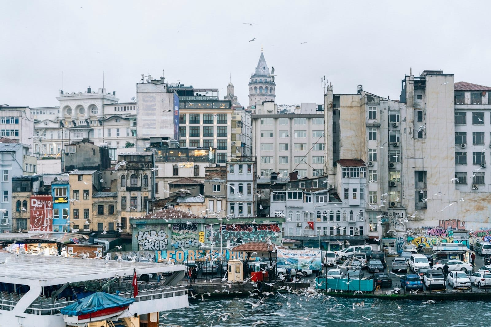 วิวริมน้ำของ Karaköy พร้อมด้วยอาคารประวัติศาสตร์ กราฟฟิตีสีสันสดใส เรือเฟอร์รี่ที่จอดเทียบท่า และหอคอย Galata ที่สูงตระหง่านเหนือทิวทัศน์เมือง Istanbul ในวันที่มีเมฆครึ้ม