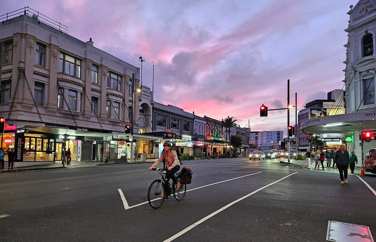 ถนน Karangahape ยามเย็น อาคารเก่า ร้านค้าสีสด ป้ายคาเฟ่ และผู้คนเดินปั่นจักรยานที่แยกคึกคักท่ามกลางท้องฟ้ายามค่ำสีจัดจ้าน