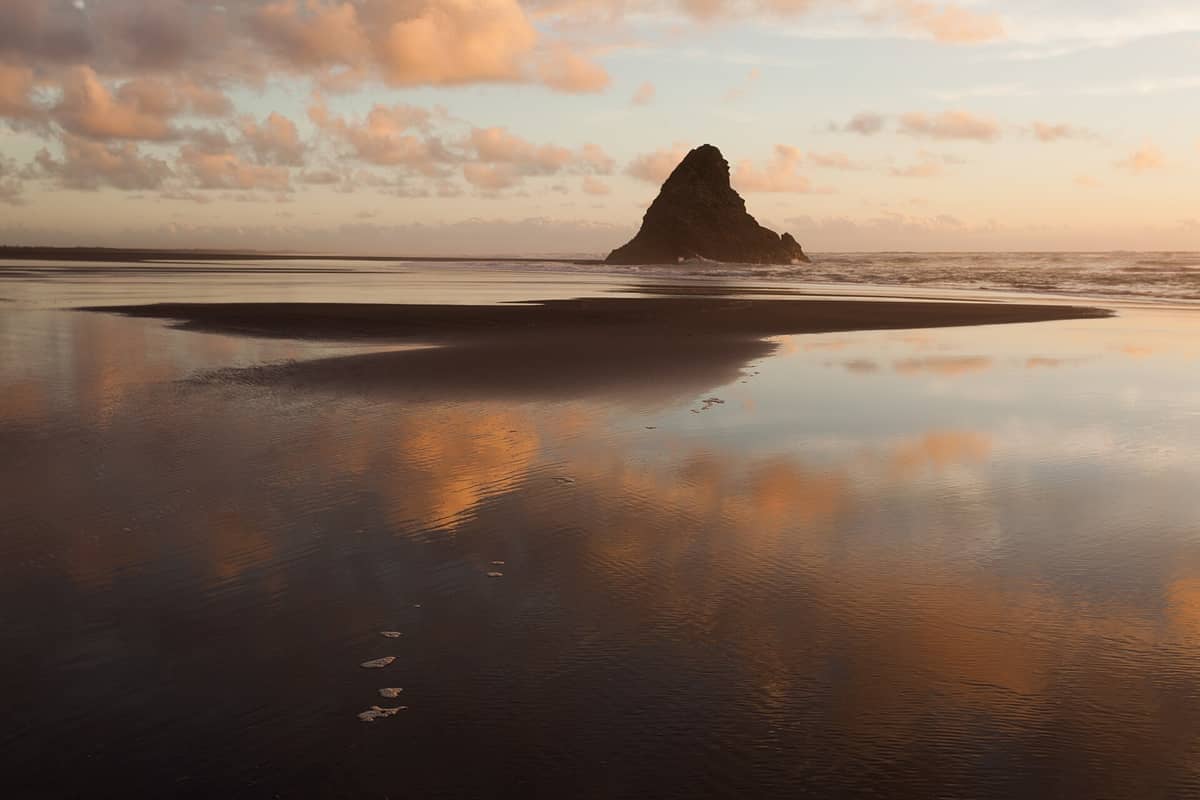 Weitläufiger Blick auf den schwarzen Sandstrand von Karekare Beach mit einer markanten Felsformation, sanften Wellen und Wolken, die sich bei Sonnenuntergang im nassen Sand spiegeln.