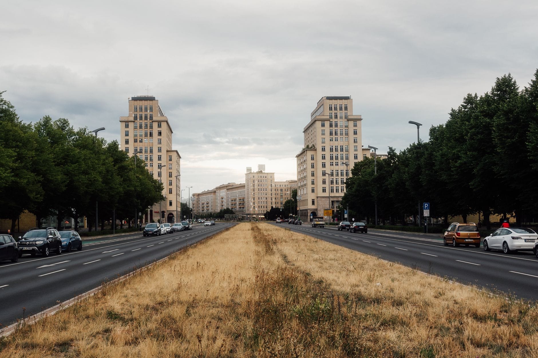 Vista del lungo viale Karl-Marx-Allee a Berlino con l'imponente architettura stalinista, fiancheggiato da torri residenziali, alberi e auto sotto un cielo nuvoloso.