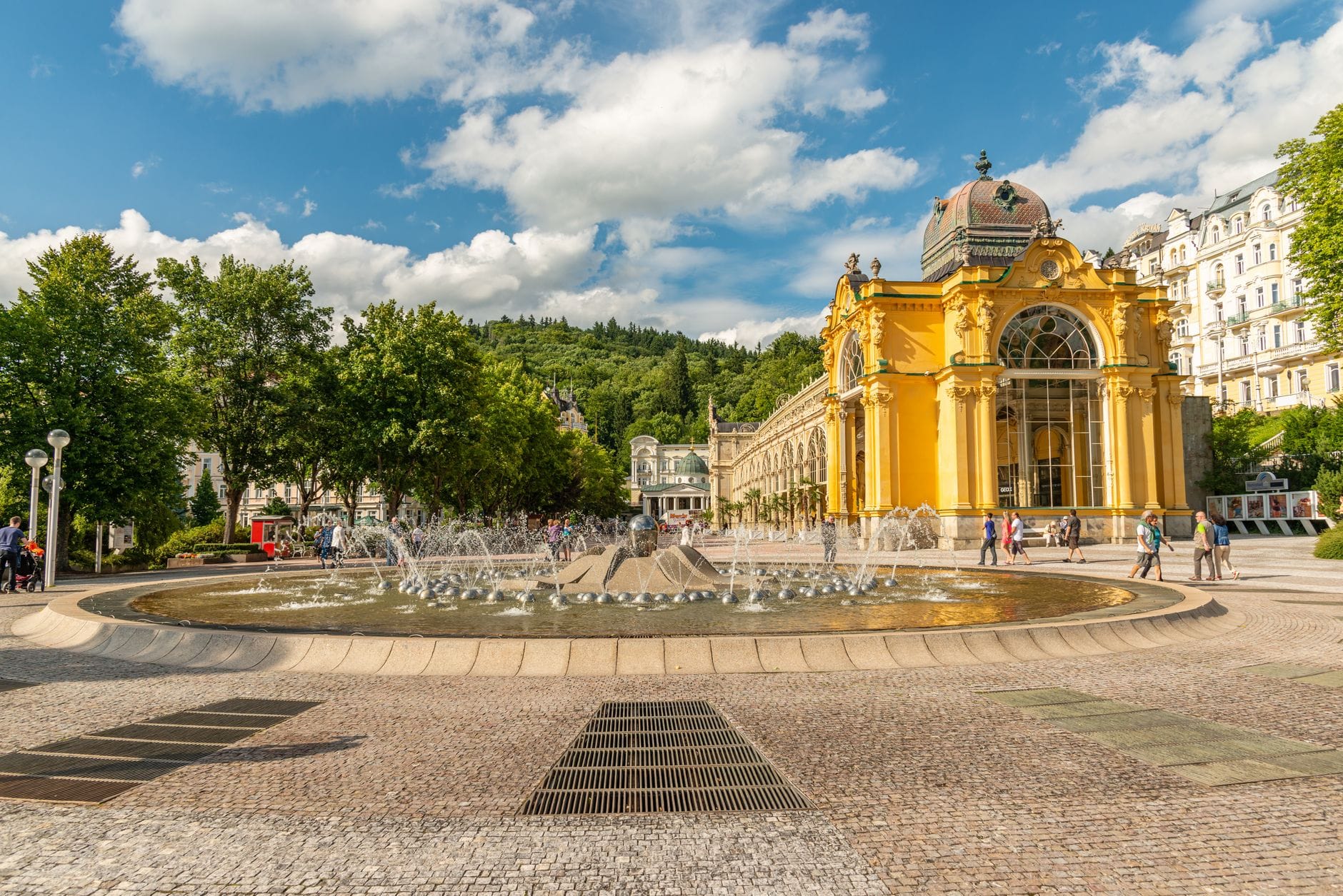 Vista panoramica dell'iconica colonnade gialla e dell'elegante edificio termale con una grande fontana sul davanti, circondati da una lussureggiante vegetazione sotto un cielo azzurro a Karlovy Vary.