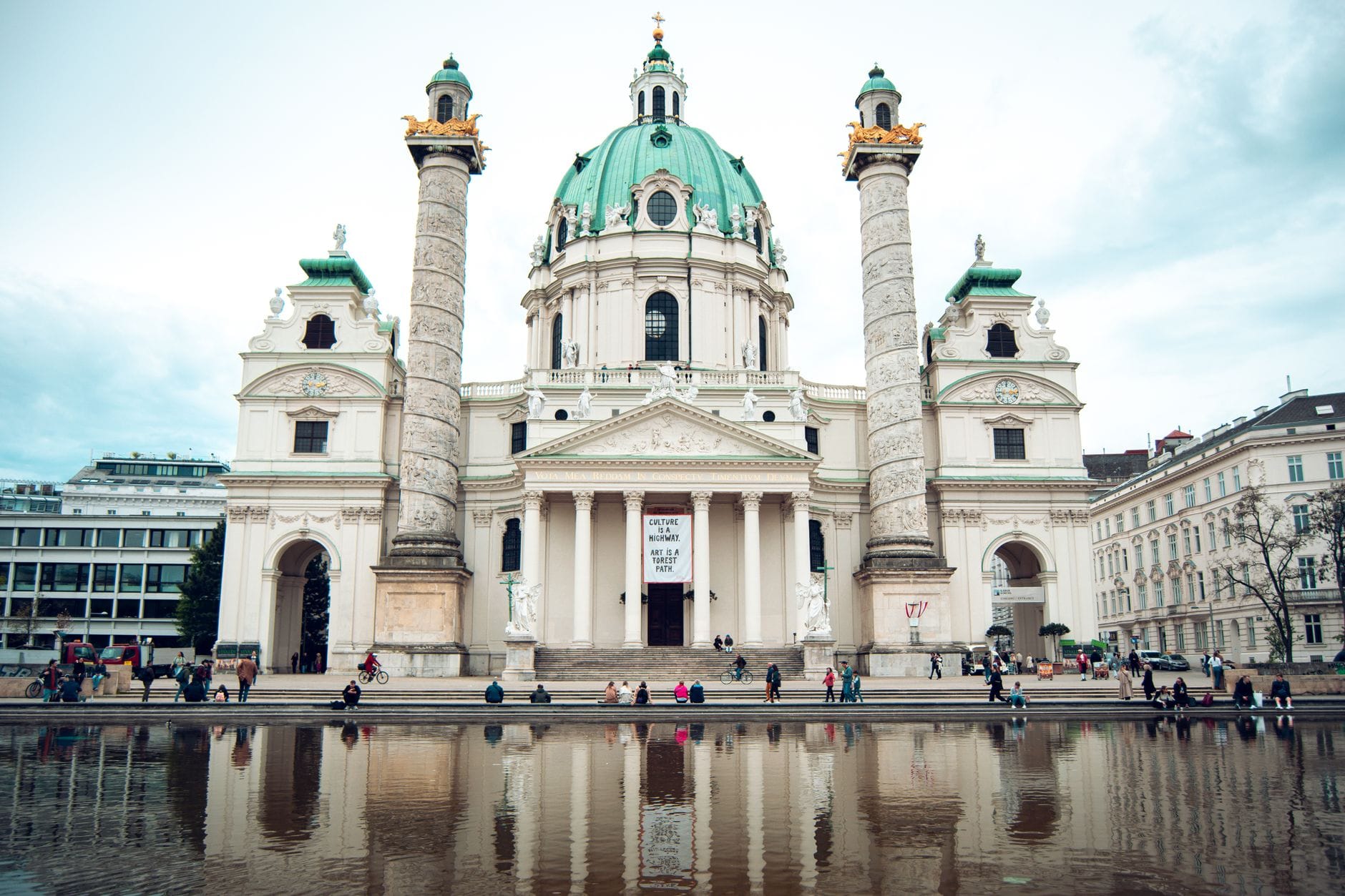 Frontansicht der Karlskirche mit Kuppel, zwei Säulen und Spiegelung im Teich, Menschen sitzen an einem bewölkten Tag rund ums Wasser.