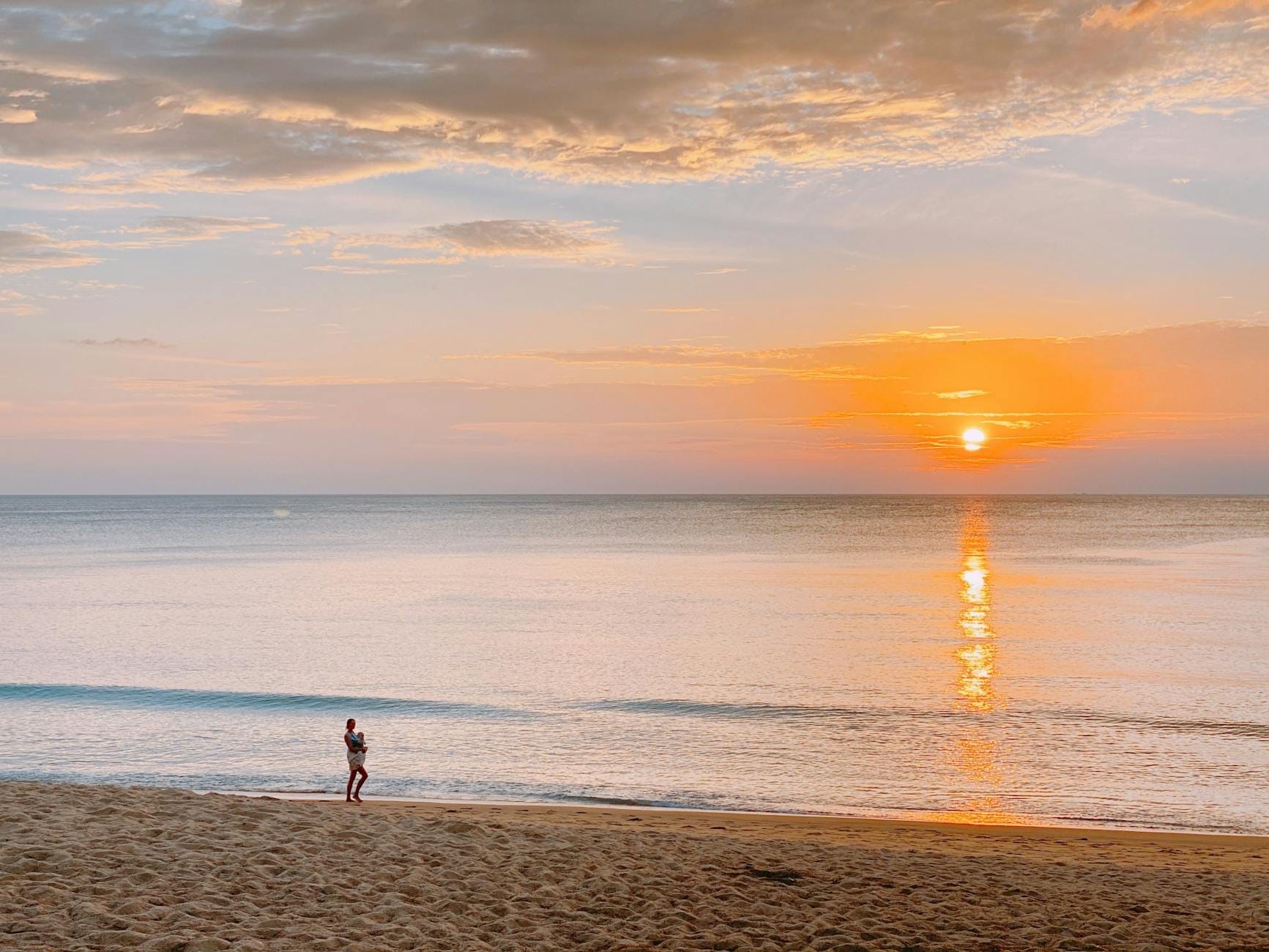 Wide sandy beach at sunset with calm waves, clear sky, and a single person walking along the shoreline, creating a peaceful and inviting atmosphere.