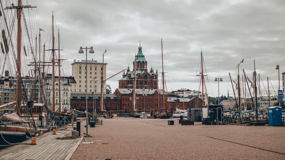 Vista da orla de Katajanokka com veleiros históricos, edifícios Art Nouveau e a Catedral Uspenski em tijolos vermelhos dominando o fundo sob um céu nublado.