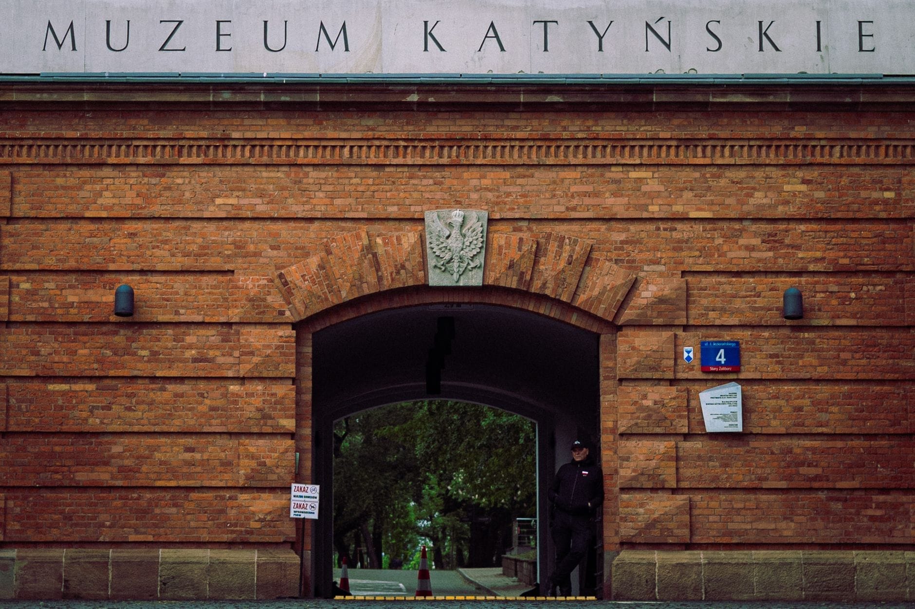 ทางเข้าพิพิธภัณฑ์กาตินในป้อมวอร์ซอซิตาเดล กำแพงอิฐสีแดง ป้าย 'Muzeum Katyńskie' เหนือประตูโค้ง และผู้คุมยืนอยู่เดี่ยว ๆ