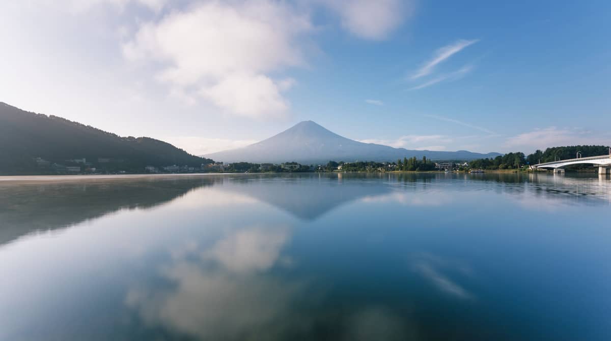 Wide view of Lake Kawaguchiko with Mount Fuji reflected on calm water and the Kawaguchiko Ohashi Bridge under a blue sky with some clouds.