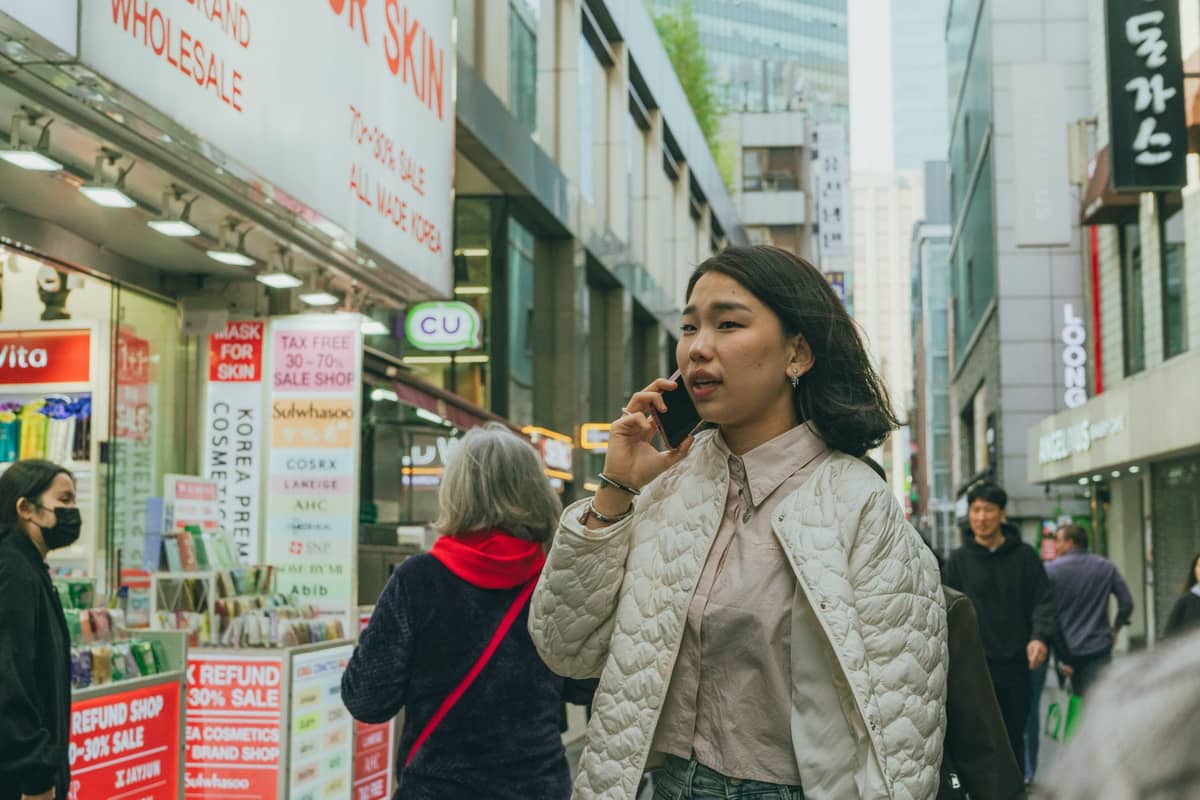 Cena de rua em Seul com pessoas caminhando por lojas de K-beauty, placas de cuidados com a pele e banners de promoções em um movimentado distrito comercial.